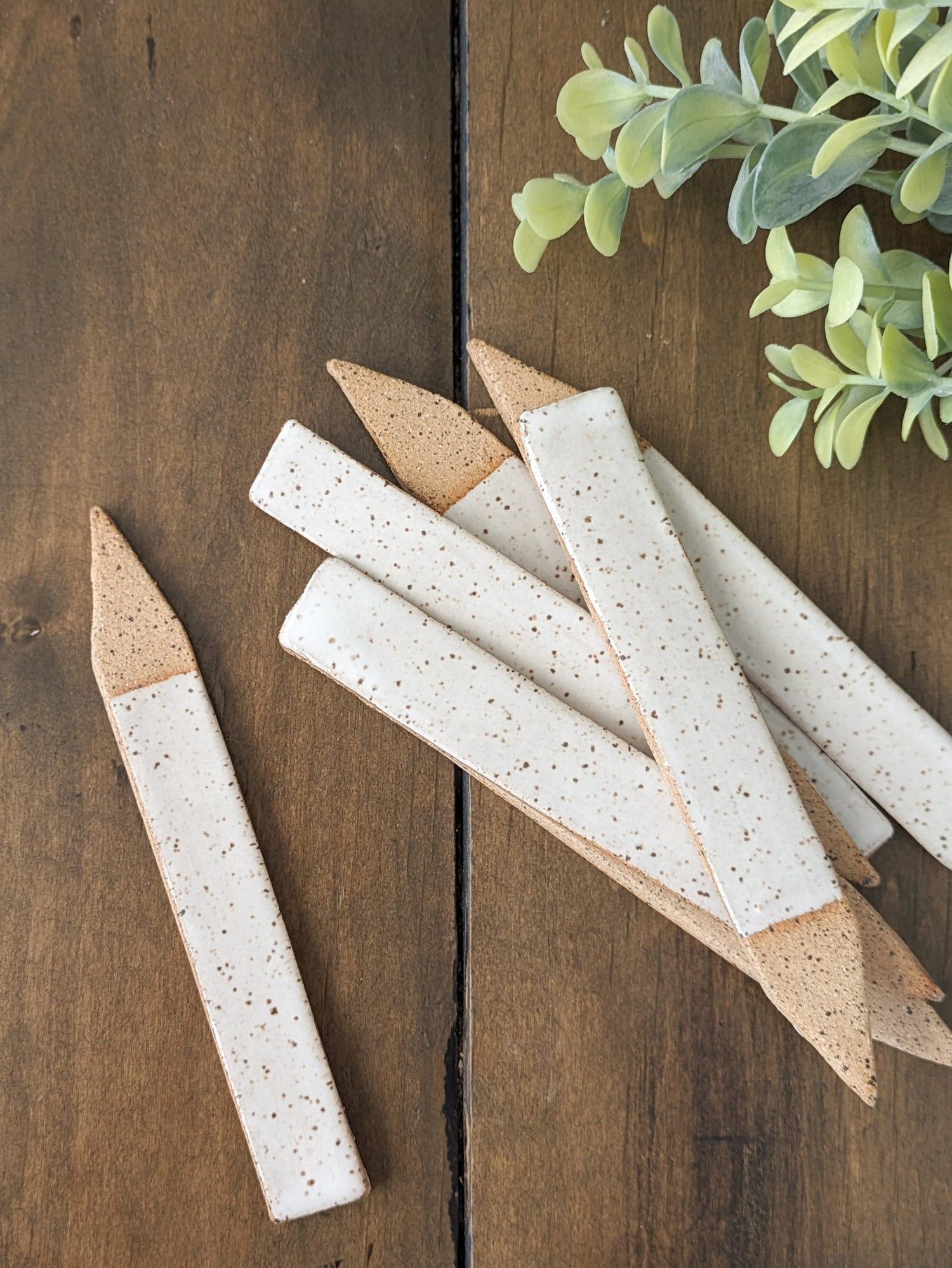 May include: A set of seven speckled, off-white ceramic plant markers with tan tips.  The markers are arranged in an overlapping stack, with one marker placed separately. The markers have a rustic, handmade appearance.  They are displayed on a dark brown wooden surface. The plant markers are ideal for labeling plants in gardens or pots.